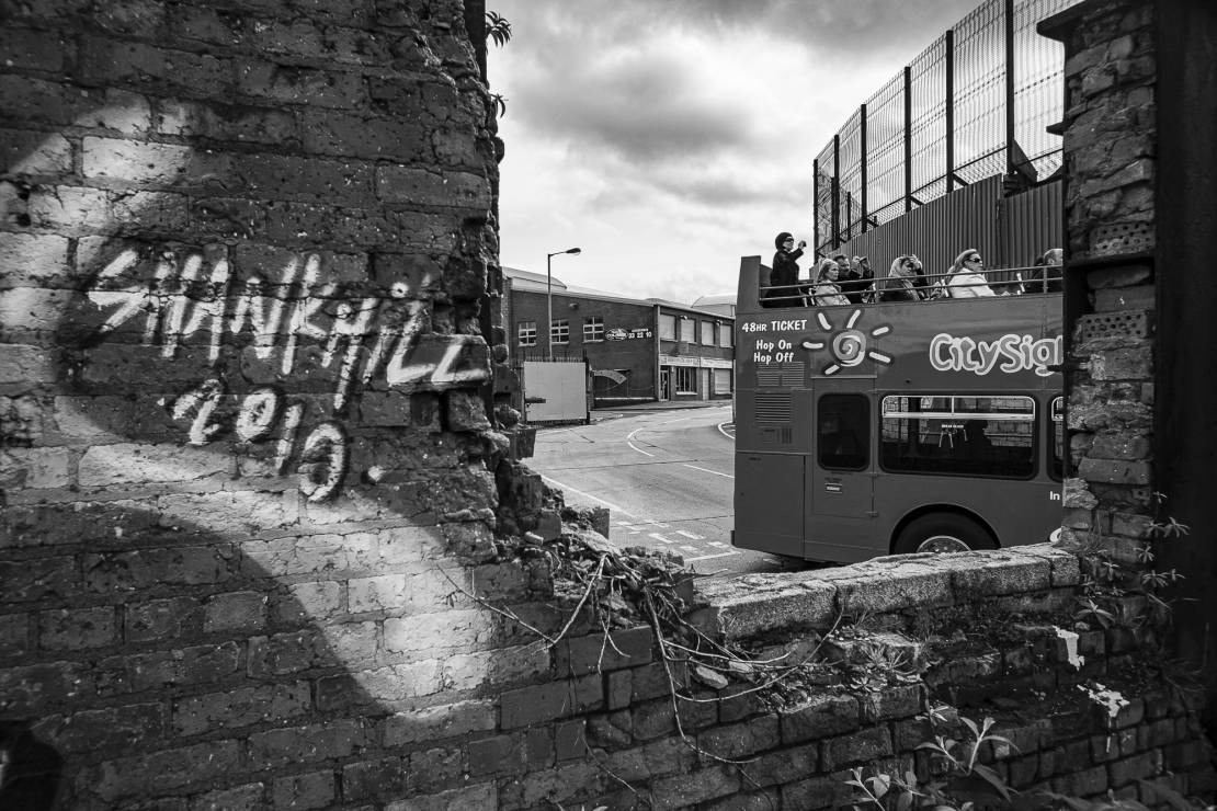 Turyści przyjeżdżający do Belfastu z dystansu oglądają podzielone miasto z piętrowych autobusów. W tle znajduje się najdłuższa „ściana pokoju” w Belfaście, oddzielająca katolików od protestantów.