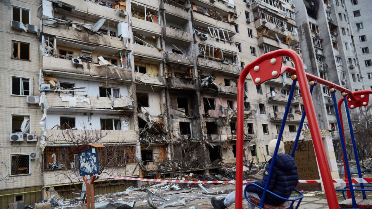 Dziecko, które buja się na huśtawce, spogląda na budynek w Kijowie zniszczony przez rakietę, fot. Pierre Crom/Getty Images